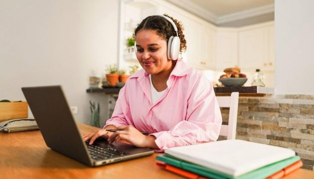 Student in pink shirt studying on a laptop with headphones and folders on the desk