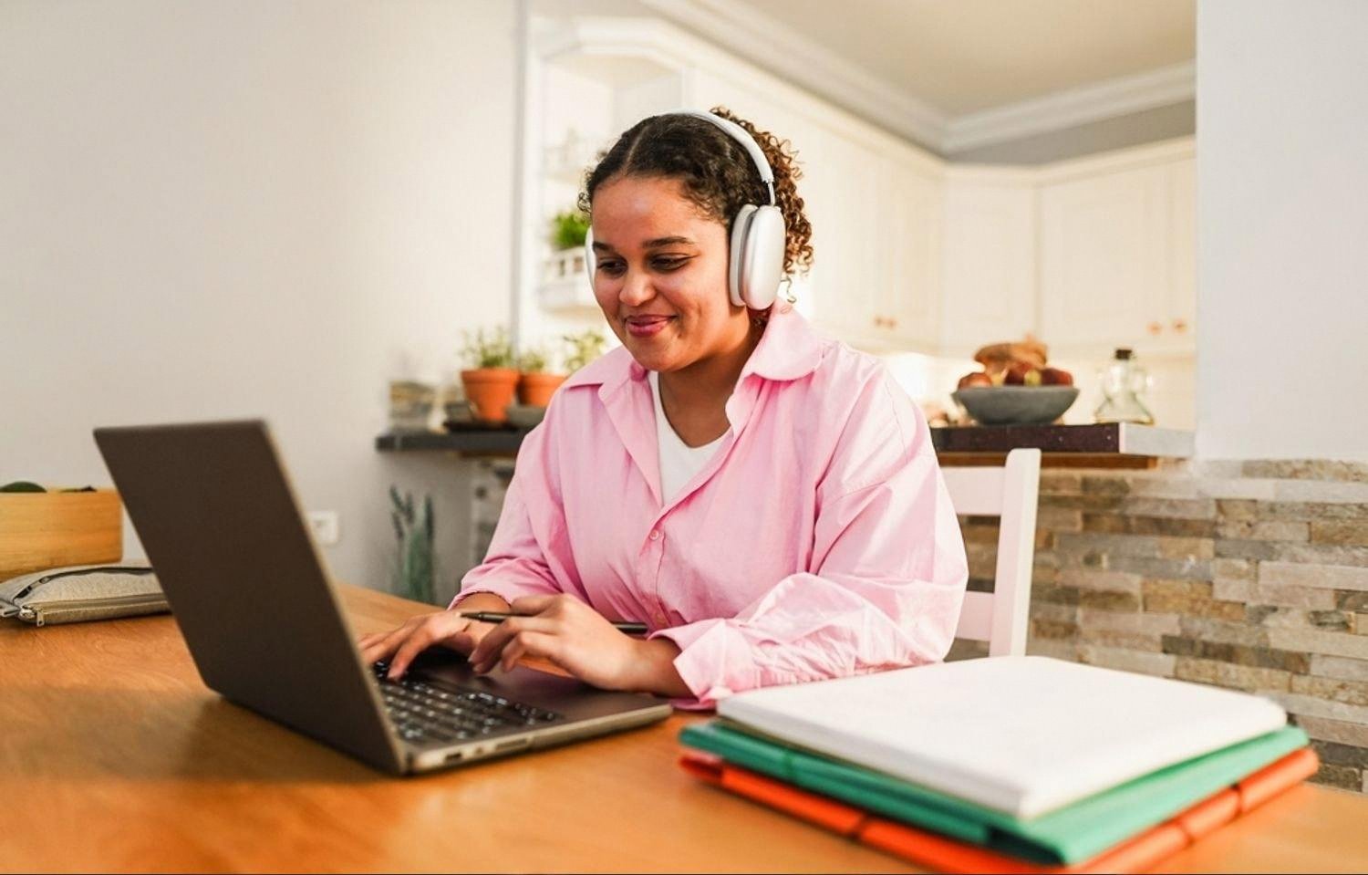 Student in pink shirt studying on a laptop with headphones and folders on the desk